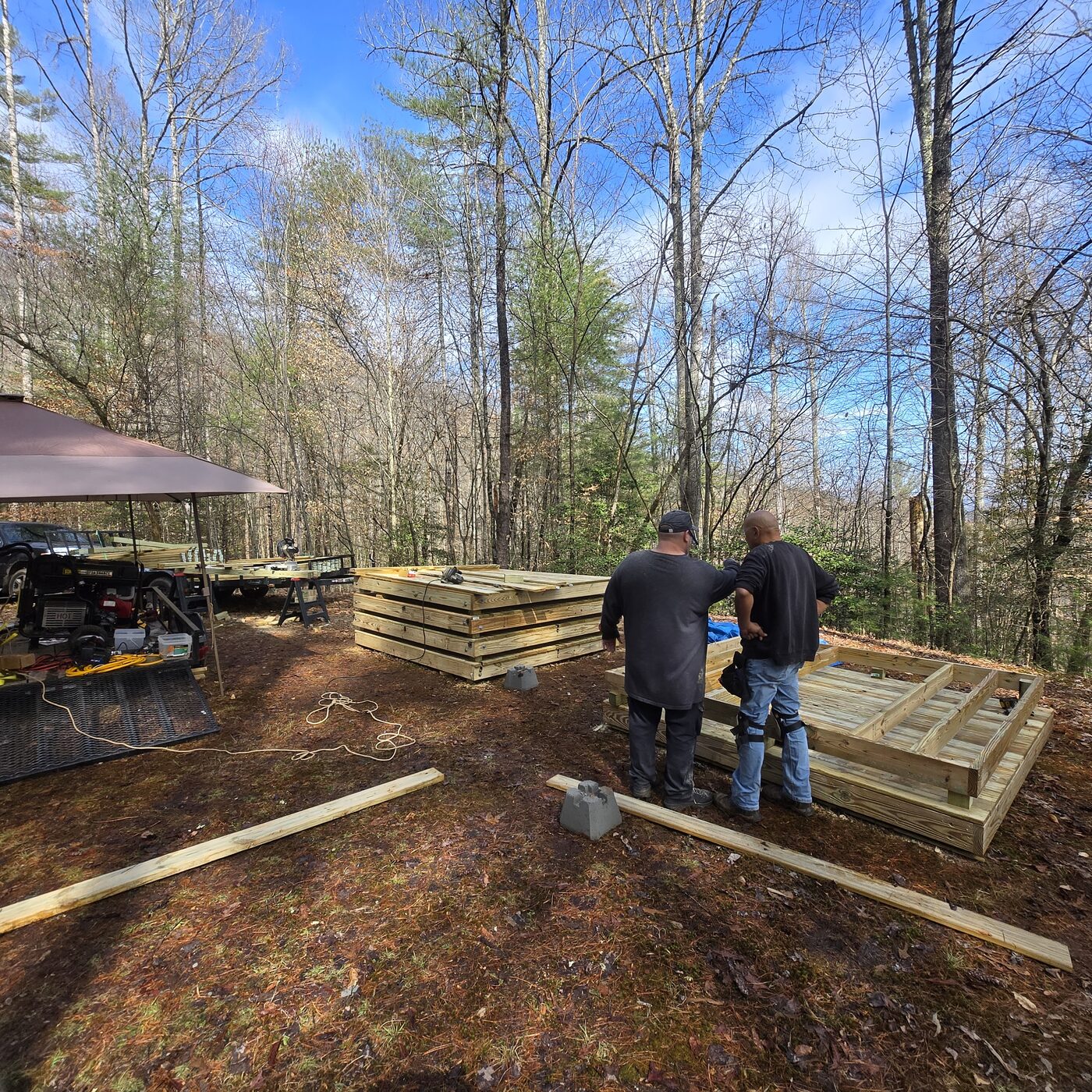 Framing tent deck panels at Cardinal Mountain Campground: a work canopy, stacked finished panels and two builders in winter woods near Franklin, NC