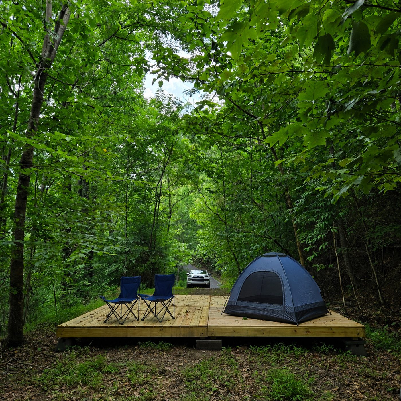 A tent and two camp chairs on the deck at Pisgah 16, Cardinal Mountain Campground near Franklin NC, with a vehicle on the track behind and trees closing in